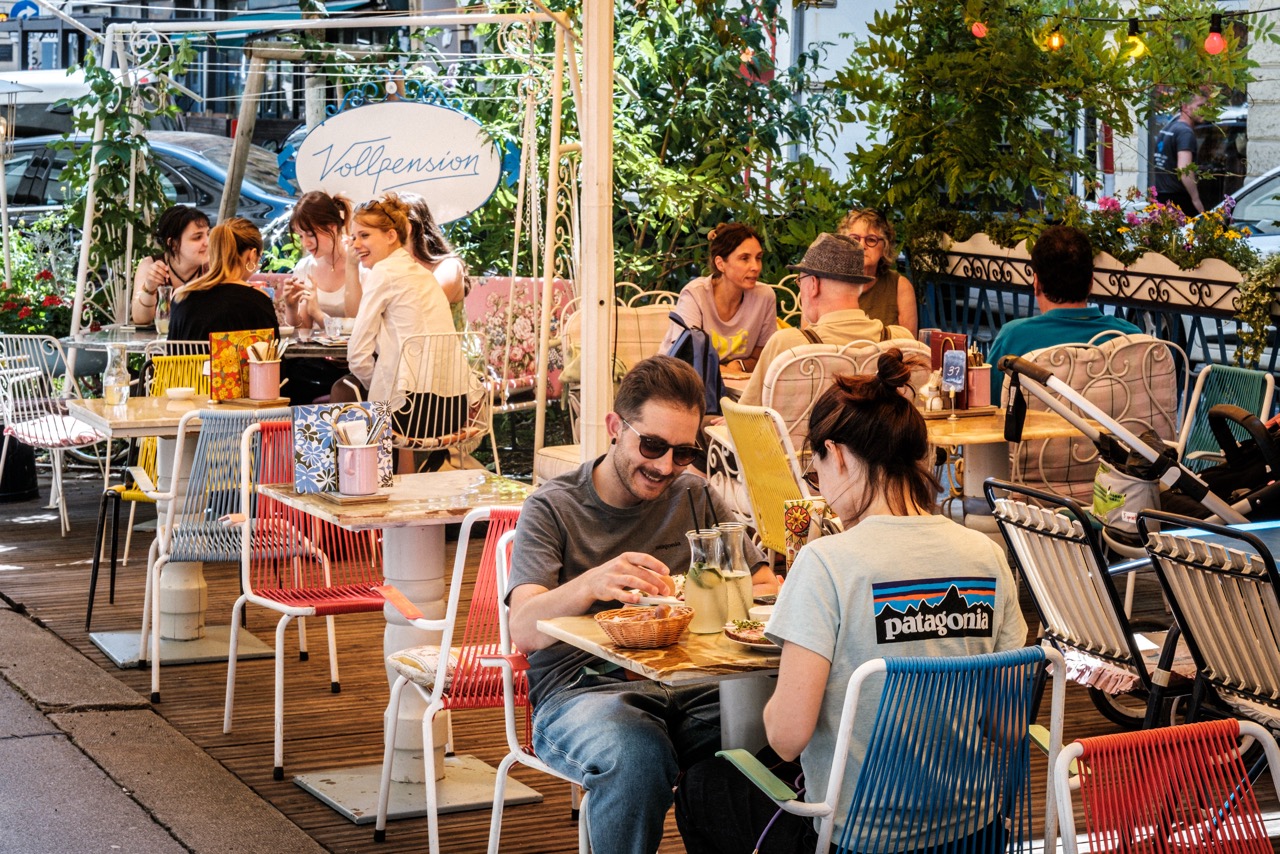 Gäste genießen Kuchen und Kaffee im sonnigen Gastgarten in Omas öffentlichem Wohnzimmer in der Vollpension Schleifmühlgasse