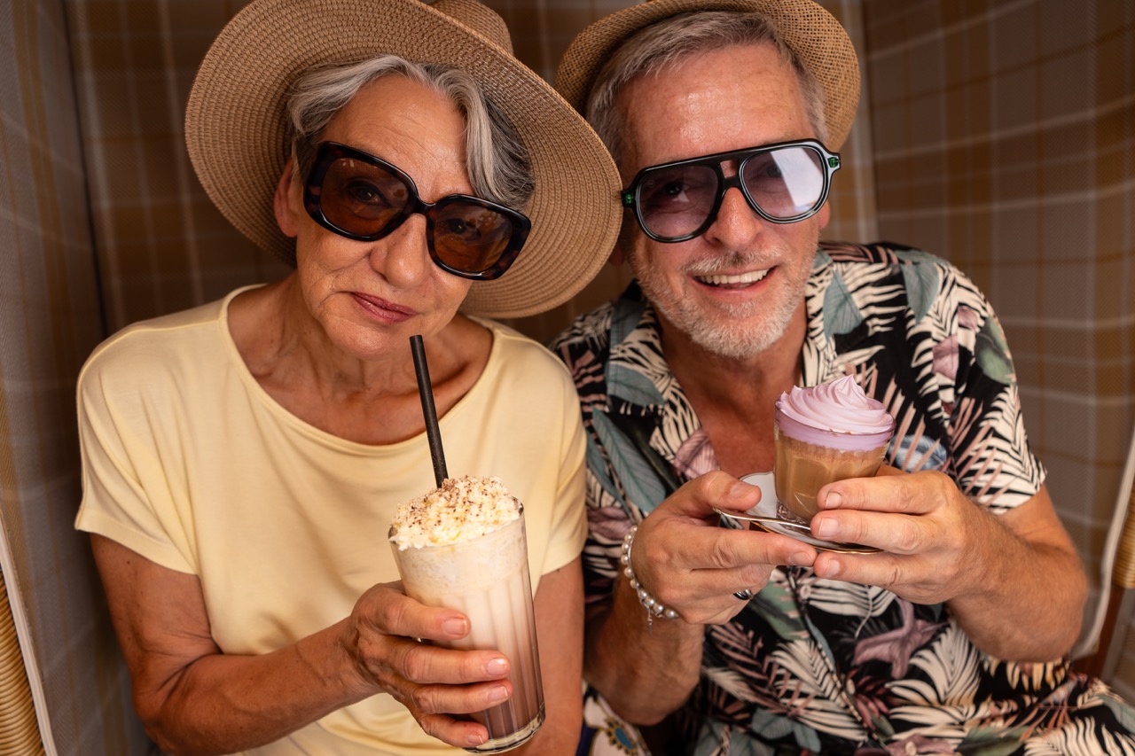 Zwei Senior:innen der Vollpension halten lächelnd einen Iced Sacher und einen Powidl-Einspänner in die Kamera